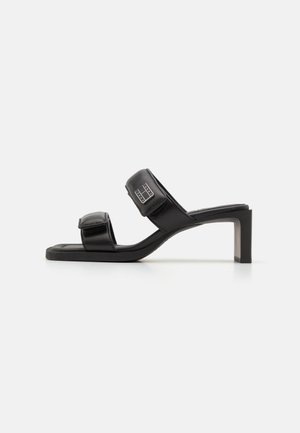 Black heeled sandals featuring two wide, adjustable straps with Velcro, a smooth leather material, and a square toe design.