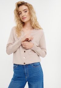 Lichtbeige cardigan met een V-hals, lange mouwen en gouden knopen; gecombineerd met blauwe high-waisted jeans. Simpele gebreide textuur.