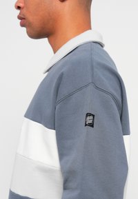Grå og hvid stribet sweatshirt med krave, lavet af blødt bomuldsstof og med et sort mærke på venstre ærme.