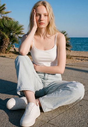 Jeune femme aux longs cheveux blonds, débardeur blanc, jean bleu clair et baskets blanches assise sur le trottoir près de la plage et des palmiers.