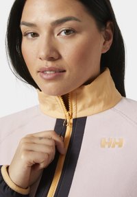 Chaqueta de forro polar con un cuerpo rosa claro, rayas navy en contraste y un cuello melocotón. Cuenta con un cierre de cremallera y un logo en el pecho.