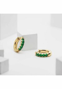 Guldöglor med gröna ädelstenar, med en slät yta och en rad av klor-fästa stenar, placerade på en neutral bakgrund.