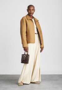 Barna bordázott dzseki, fehér felső, krém színű, széles szárú nadrág és mintás barna kézi táska. A modellen fonott szandál van, a kezeit a csípőjére tette.