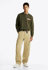 Sudadera verde oliva con parche de logo, combinada con pantalones cargo beige que tienen múltiples bolsillos y un cinturón negro, llevada con zapatillas blancas.