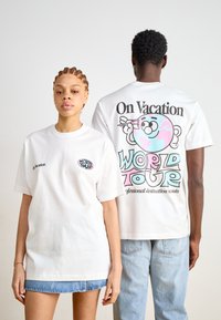 T-shirts brancos em algodão com designs gráficos. A parte da frente apresenta "De Férias" e um pequeno logótipo colorido; a parte de trás apresenta um gráfico maior com "TOUR MUNDIAL."