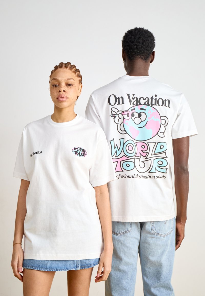 T-shirts brancos em algodão com designs gráficos. A parte da frente apresenta "De Férias" e um pequeno logótipo colorido; a parte de trás apresenta um gráfico maior com "TOUR MUNDIAL."