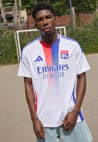 Bílý fotbalový dres s červenými a modrými vertikálními pruhy, s nápisem "Emirates Fly Better" a logem Olympique Lyonnais, krátké rukávy.