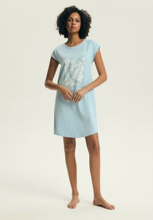 Kobieta o kręconych włosach, ubrana w jasnoniebieską, bezrękawnikową sukienkę z botanicznym nadrukiem, stojąca boso na neutralnym tle.