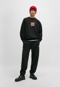 Felpa nera con logo bianco e rosso, abbinata a pantaloni della tuta neri. Berretto lavorato a maglia rosso con "HUGO" in nero. Stivali neri con lacci.