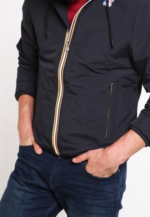 Mann trägt eine marineblaue Jacke mit Reißverschluss und gelben sowie weißen Akzenten, rotes Hemd darunter, Hände in den Taschen, blaue Jeans.