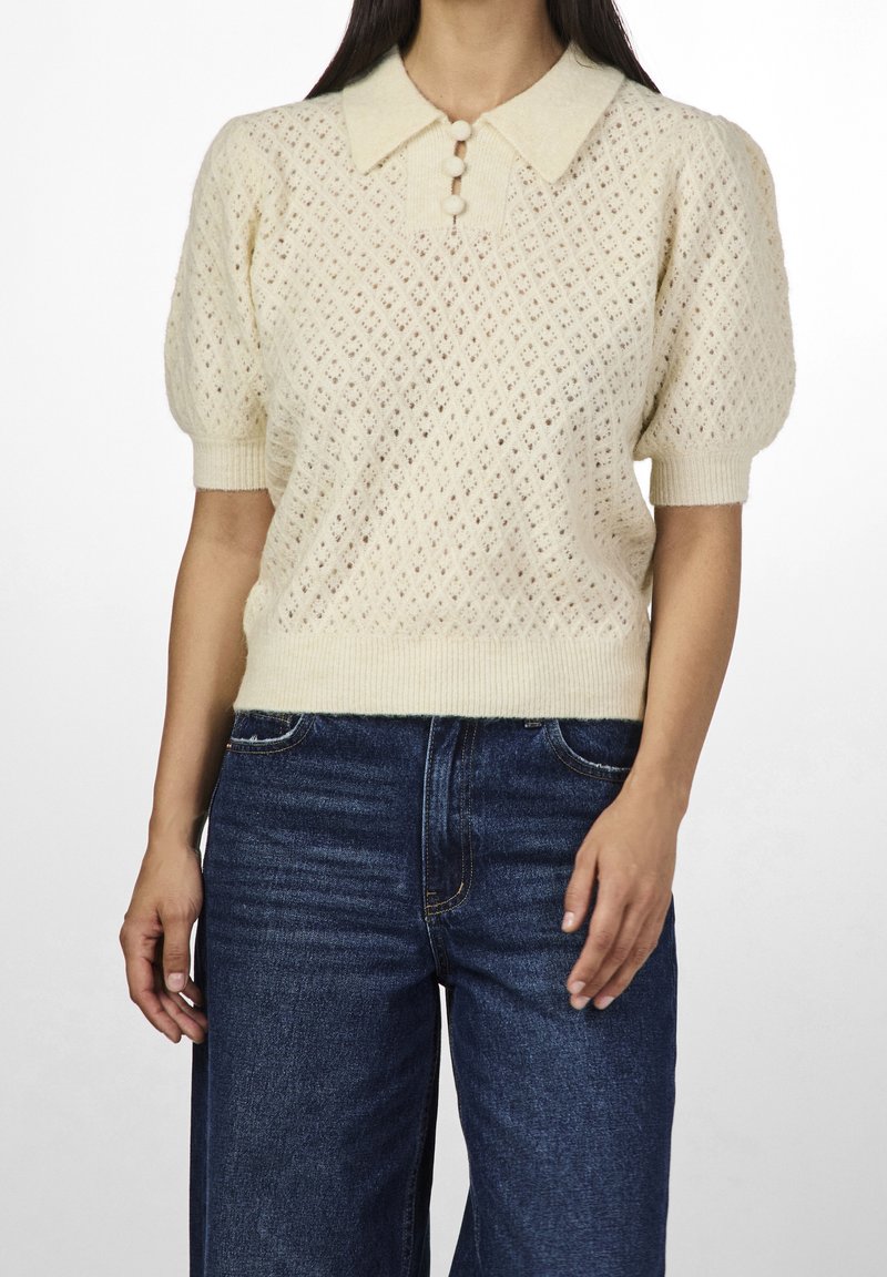 Maglione lavorato a maglia color crema, a maniche corte, con un motivo a rombi, colletto pieghevole e patta con tre bottoni; abbinato a jeans blu scuro.