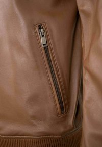 Braune Lederjacke mit einer seitlichen Reißverschlusstasche und geripptem Saum. Die Textur ist glatt mit einer matten Oberfläche.