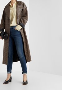 Abrigo largo a cuadros marrón sobre una camisa amarillo claro, combinado con jeans ajustados azul oscuro y zapatos de tacón negros, sosteniendo un bolso de mano negro.