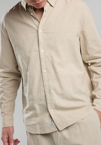 Camicia di lino beige con maniche lunghe, colletto a scialle, bottoni sul davanti e spacchi laterali, caratterizzata da un'etichetta a marchio tessuta in basso.