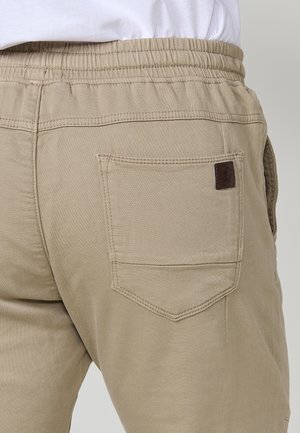 Tuvumā redzami beigē krāsas elastīgi jostas bikšu, ar aizmugurējo kabatu un nelielu iestiprinātu ādas zīmola pazīmi kabatas stūrī.