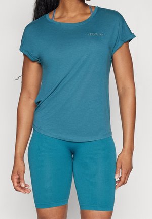 Kvinde iført teal kortærmet skjorte med "ONLY PLAY" logo og matchende ribstrikkede cykelshorts mod en ensfarvet baggrund.