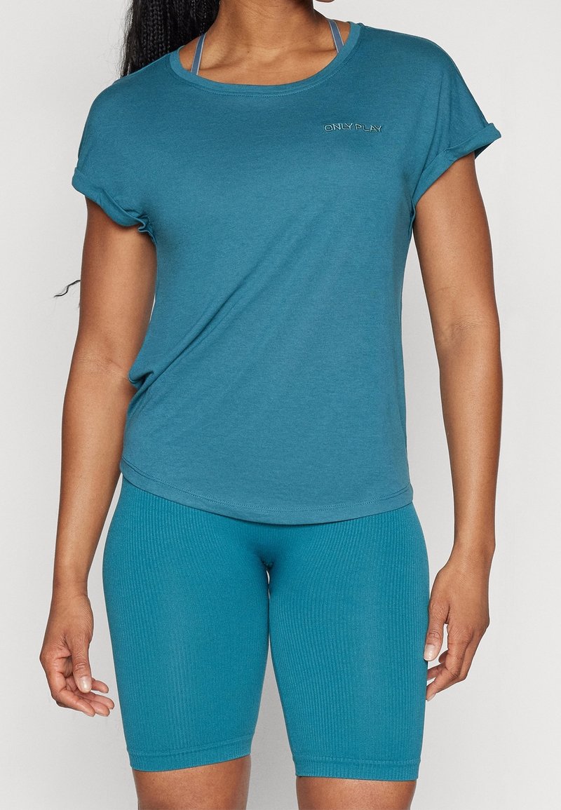 Femme portant un t-shirt à manches courtes bleu sarcelle avec le logo "ONLY PLAY" et un short de cycliste côtelé assorti sur un fond uni.
