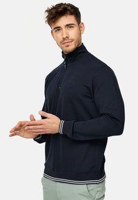Mornarica pulover s patentnim zatvaranjem i visokim ovratnikom, ribanim manžetama i donjim rubom s bijelim prugama te malim utisnutim logom na prsima.