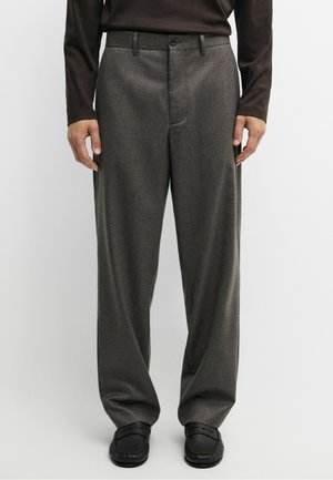 Mand iført mørkebrun bluse med lange ærmer, løstsiddende grå bukser og sorte loafers står mod en ensfarvet lys baggrund.