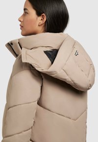 Beige Daunenjacke mit hohem Kragen, strukturiertem Nähen und schwarzem Innenfutter. Verfügt über ein Logo-Akzent auf dem Rücken und einen Reißverschluss.