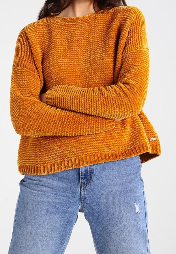 Gestrickter oranger Pullover mit strukturiertem Muster und gerippten Bündchen, kombiniert mit blauen, ausgewaschenen Jeans. Der Pullover hat eine lockere Passform.