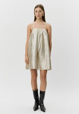 Gouden, mouwloze jurk van satijnachtige stof met een geplooid ontwerp, uitlopend vanaf het lijfje. Draagt met puntige zwarte laarzen van krokodillenleer.