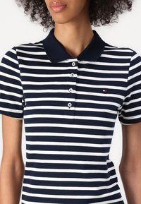 Femme portant un polo à rayures horizontales bleu marine et blanc, manches courtes et patte boutonnée, coupé au niveau du menton et du milieu du torse.