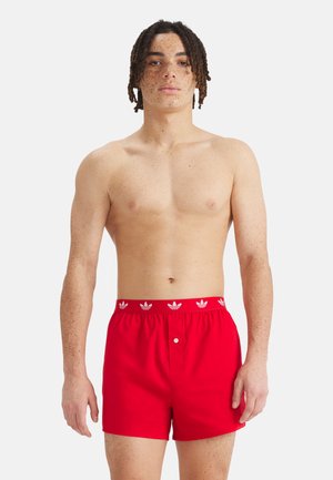 Mladý muž s pihami v červených boxerkách značky Adidas stojí před jednoduchým bílým pozadím.