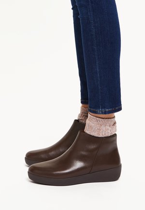 Bottes de cheville en cuir marron avec une texture lisse, un bout rond et un talon compensé bas, associées à des chaussettes en tricot roses et un jean sombre.