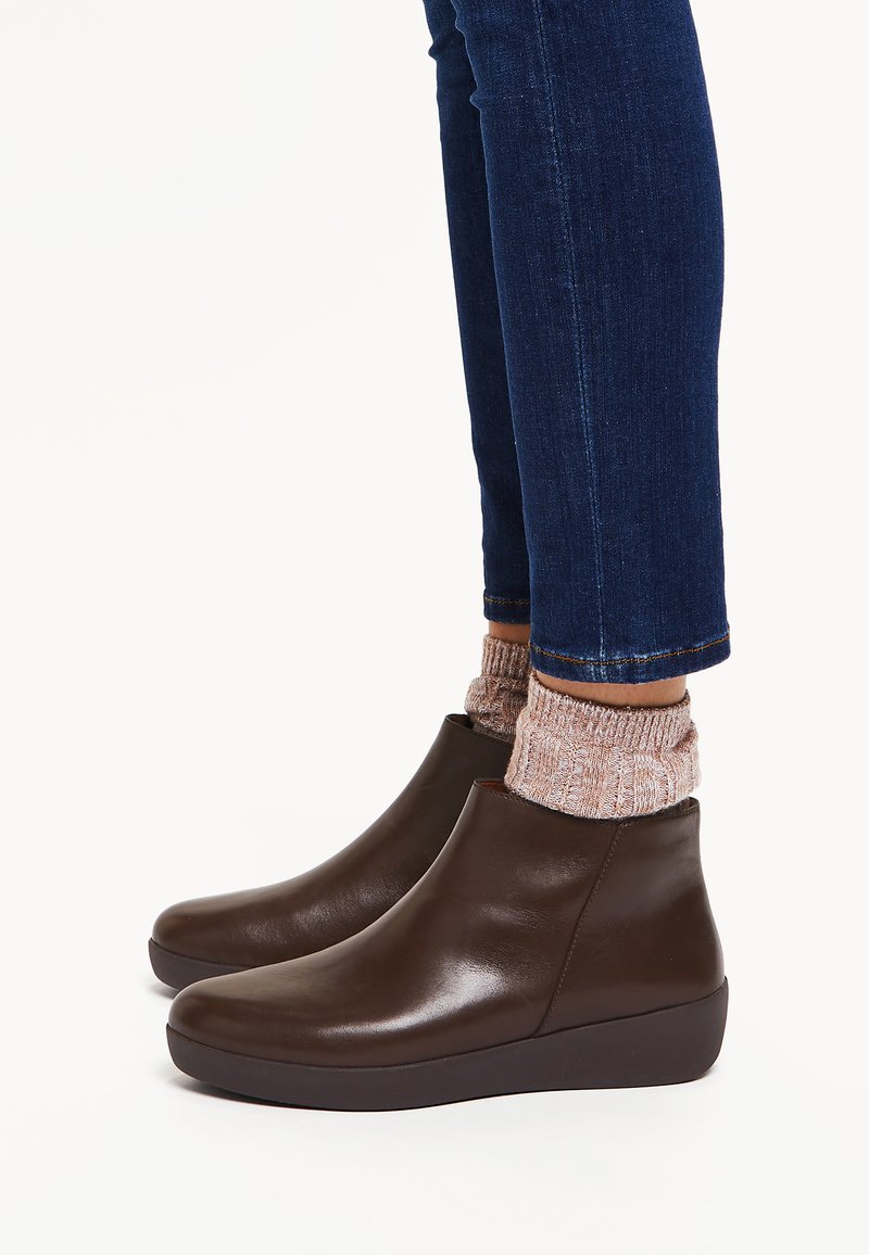 Stivali alla caviglia in pelle marrone con una texture liscia, punta tonda e tacco a zeppa basso, abbinati a calze rosa lavorate a maglia e jeans scuri.