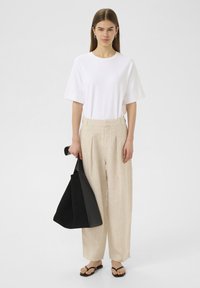 Witte t-shirt met korte mouwen, gecombineerd met beige linnen wijde broek. Zwarte totebag in de hand en zwarte teenslippers aan de voeten.