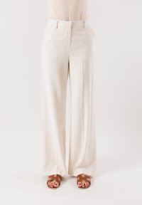 Pantaloni larghi crema con una texture liscia, pieghe davanti, passanti per cintura e un taglio su misura, abbinati a sandali piatti marroni.