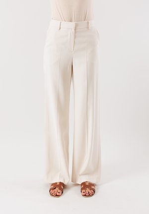 Pantaloni larghi crema con una texture liscia, pieghe davanti, passanti per cintura e un taglio su misura, abbinati a sandali piatti marroni.