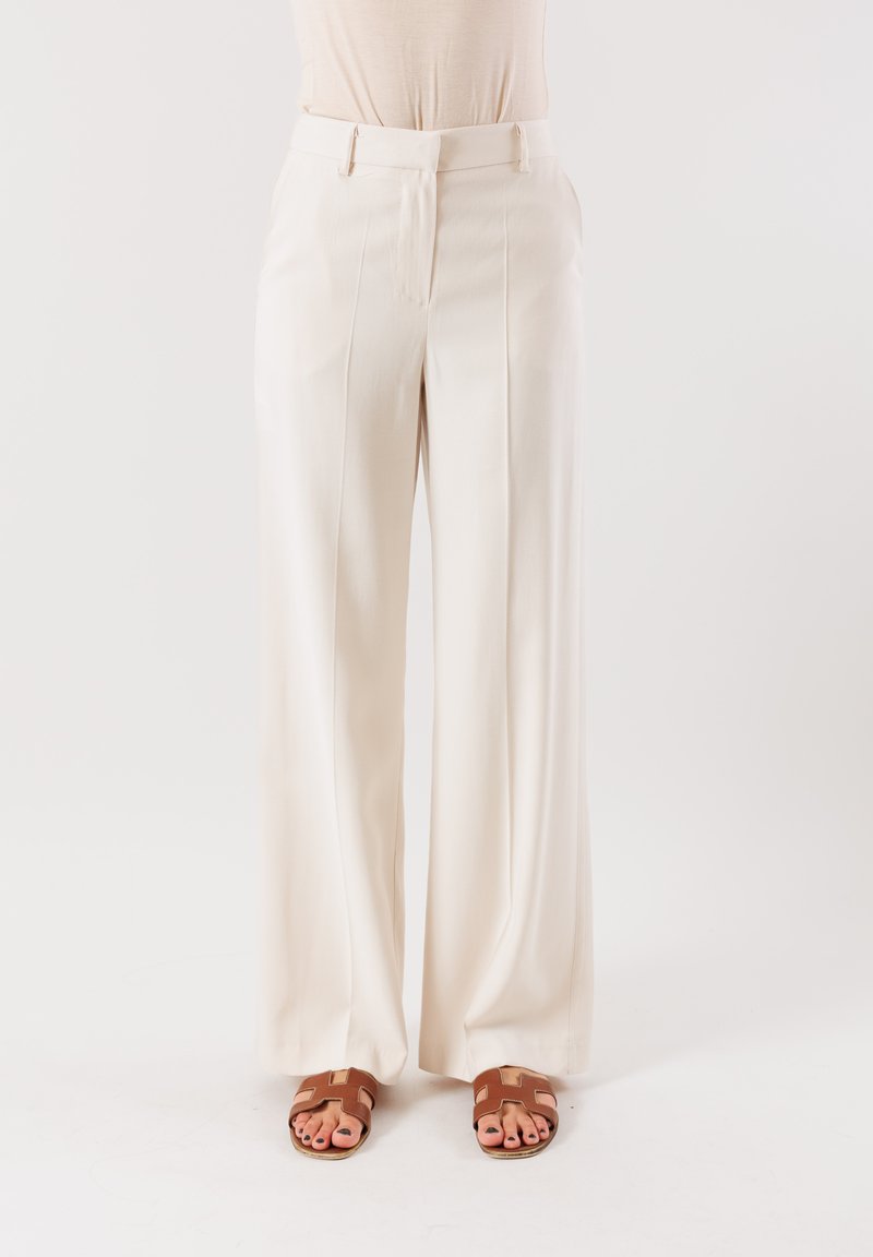 Pantaloni larghi crema con una texture liscia, pieghe davanti, passanti per cintura e un taglio su misura, abbinati a sandali piatti marroni.