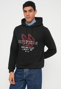 Čierna mikina s kapucňou s potlačou na prednej strane obsahujúcou červený a biely text. Nastaviteľná kapucňa so šnúrkami, rebrované manžety a mäkký materiál s uvoľneným strihom.