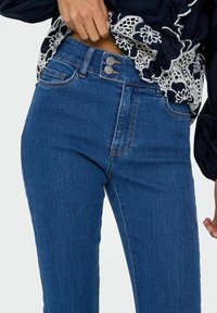 Los jeans de mezclilla azul presentan una cintura alta con cierre de doble botón, bolsillos laterales y una textura suave con un sutil desvanecimiento a lo largo de las piernas.