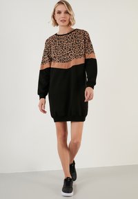Schwarzes Sweatshirtkleid mit einem Leopardenmuster im oberen Bereich und einem einfarbigen schwarzen unteren Teil. Lange Ärmel und Rippbündchen. Getragen mit schwarzen Sneakers.