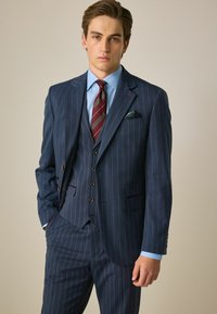 Costume rayé marine avec accents rouges, gilet croisé, pantalons assortis et chemise bleu clair. Comprend un mouchoir de poche.