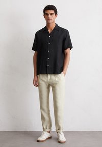 Schwarze Leinen-Hemden mit kurzen Ärmeln und Knöpfen, kombiniert mit hellbeigen schmalen Hosen. Die Schuhe bestehen aus weißen Sneakers mit einer hellbraunen Sohle.