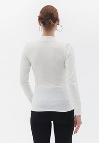 Langærmet hvid top i en tætsiddende design, lavet af blødt, let stof. Har en høj hals og glat tekstur. Bagudsigten vises.