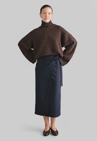 Brązowy sweter z dzianiny z golfem i dużymi rękawami, zestawiony z ciemnoniebieską kopertową spódnicą. Modelka ma na sobie ciemnobrązowe baleriny. Na szarym tle.