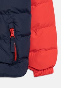 Kurtka puchowa z granatowym korpusem i czerwonymi rękawami, z widoczną kieszenią na zamek błyskawiczny oraz elastycznymi mankietami dla lepszego dopasowania.