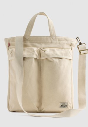 Sac en toile beige avec deux poignées courtes, une bandoulière longue réglable et deux poches avant. Présente une étiquette avec des détails de logo.