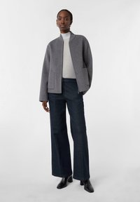 Grau melierter Wollmix-Jacke mit kragenlosem Design, vorderen Taschen, kombiniert mit dunkelblauen, weit geschnittenen Jeans und schwarzen Knöchelstiefeln.