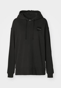 Svart hoodie i mjukt tyg med kängurufickor, dragsko i huvan och en liten märkespatch i det övre vänstra hörnet.