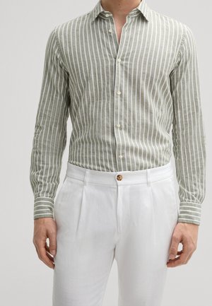 Mężczyzna ubrany w jasnozieloną i białą koszulę z pionowymi paskami z długim rękawem, włożoną do białych plisowanych spodni z brązowym guzikiem.