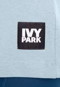 Camiseta de color azul claro con una tela texturizada. Presenta una etiqueta rectangular negra con el texto blanco "IVY PARK" en el frente.