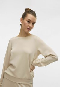 Maglietta beige a maniche lunghe con scollo rotondo e maniche con polsini. Tessuto liscio con una vestibilità rilassata. Abbinata a un capo inferiore coordinato.