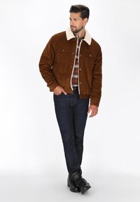 Bruin corduroy jasje met een crèmekleurige nepbont kraag, knoopsluiting, twee borstzakken, gecombineerd met blauwe jeans en zwarte laarzen.