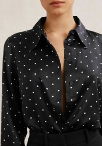 Mustast satiinist pluus valgete täpiliste täppidega, millel on terava krae, kinnituseks nööbid ja sügav V-kaelus. Sile tekstuur.